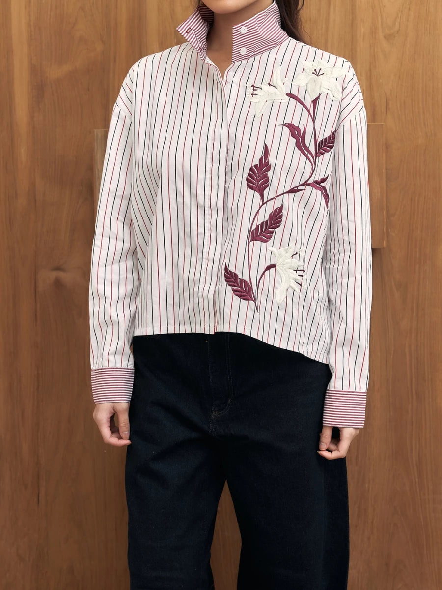 Person wearing a white shirt with red stripes and floral embroidery by Studio Tui against a wooden background