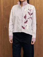 Person wearing a white shirt with red stripes and floral embroidery by Studio Tui against a wooden background