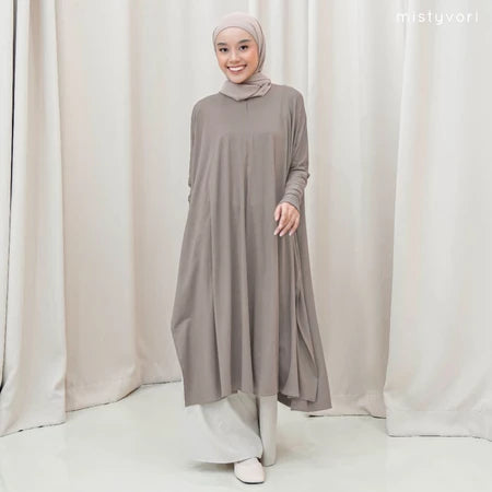 Woman wearing a long, light gray tunic by Mistyvori with a matching hijab against a white curtain background.