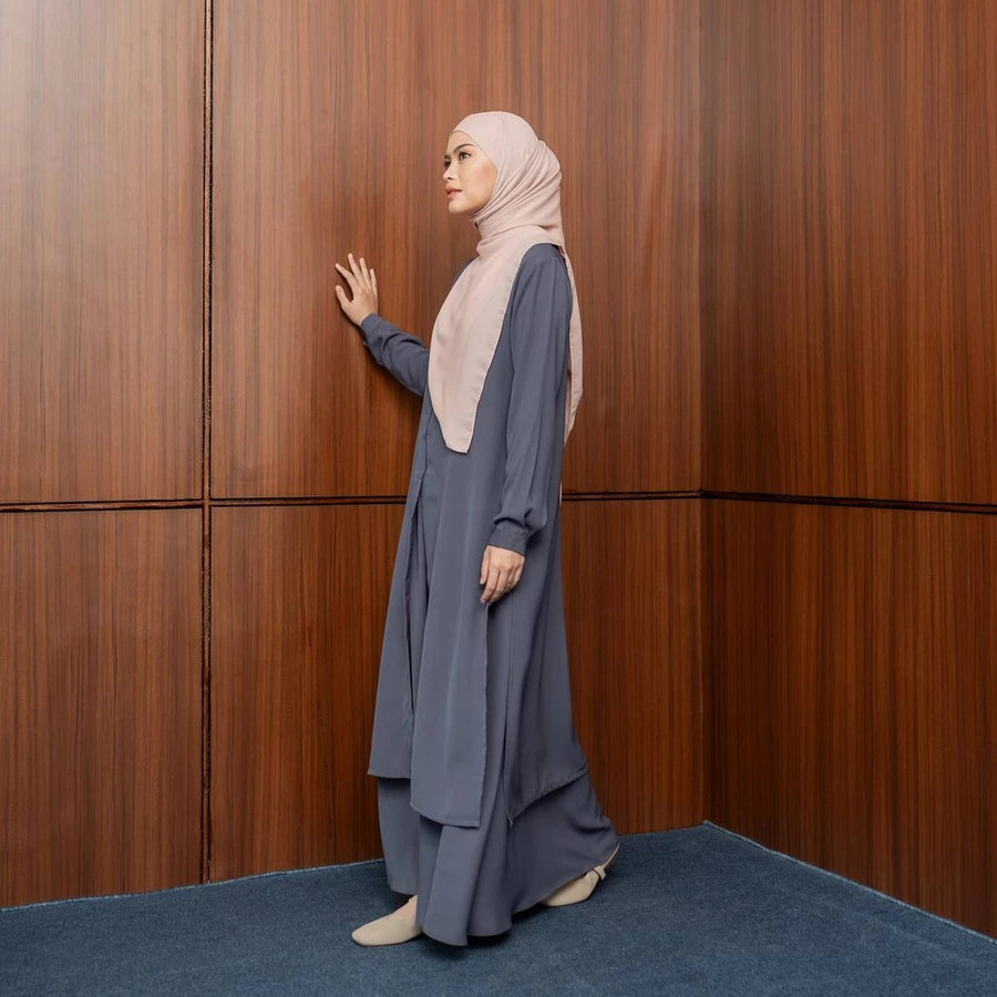 Woman in a long, dark grey tunic and beige hijab standing against a wooden paneled wall.

Seorang wanita mengenakan tunik panjang berwarna abu-abu gelap dan hijab berwarna beige, berdiri di depan dinding berpanel kayu.
