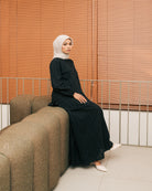 Woman in a black tunic from Atala Scarves and hijab sitting on a brown couch with wooden blinds in the background