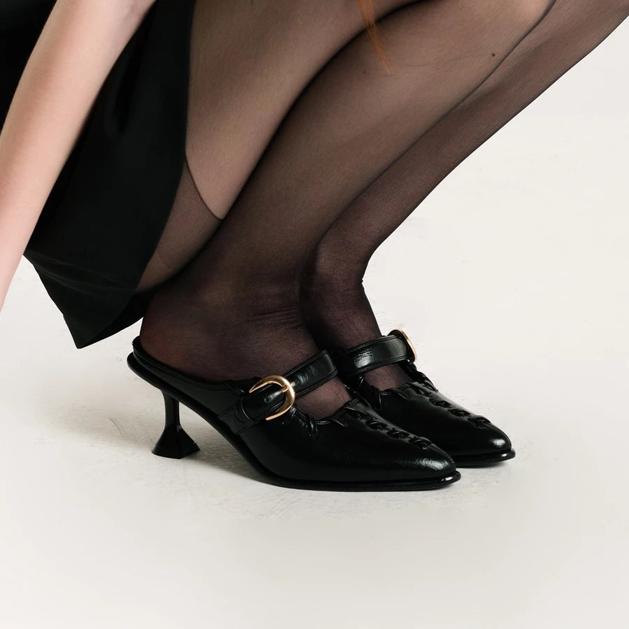 Black high-heeled shoes by Cekhas with gold buckles worn by a person in black stockings on a light background