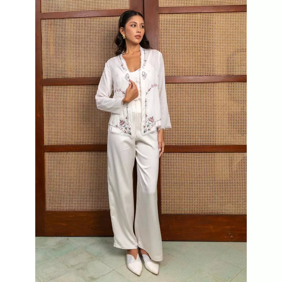 Woman in a white top with floral embroidery and white pants by Kartinis Label standing against a wooden panel background