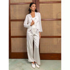 Woman in a white top with floral embroidery and white pants by Kartinis Label standing against a wooden panel background