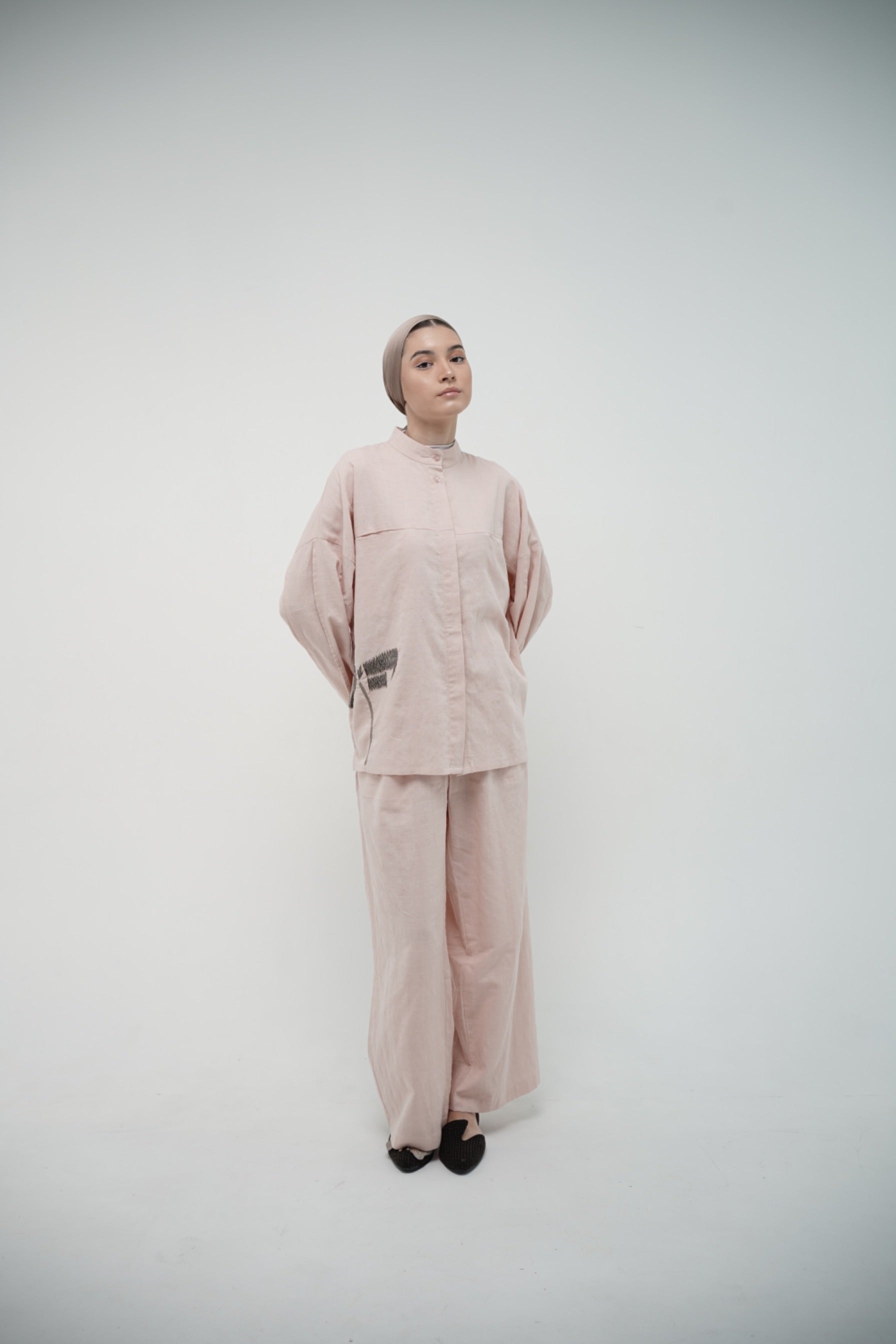 A person standing in a studio setting, wearing a pink linen top and matching pants by Glashka with a round neckline and long sleeves.