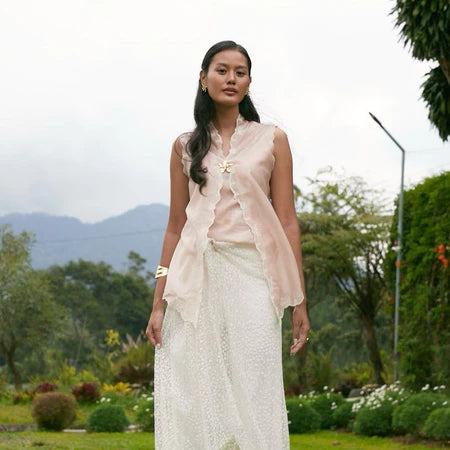 Woman in a light pink sleeveless kebaya by Kartinis Label and white skirt standing outdoors with greenery and mountains in the background
