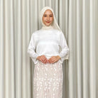 Woman wearing a white top by Kartinis Label with a hijab against a plain background