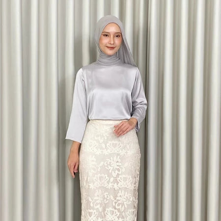 Woman wearing a light gray hijab, light gray top by Kartinis Label, and white lace skirt against a striped curtain background.