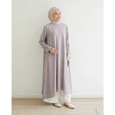 Seorang wanita memakai tunik berlengan panjang berwarna lavender, hijab, dan celana panjang.