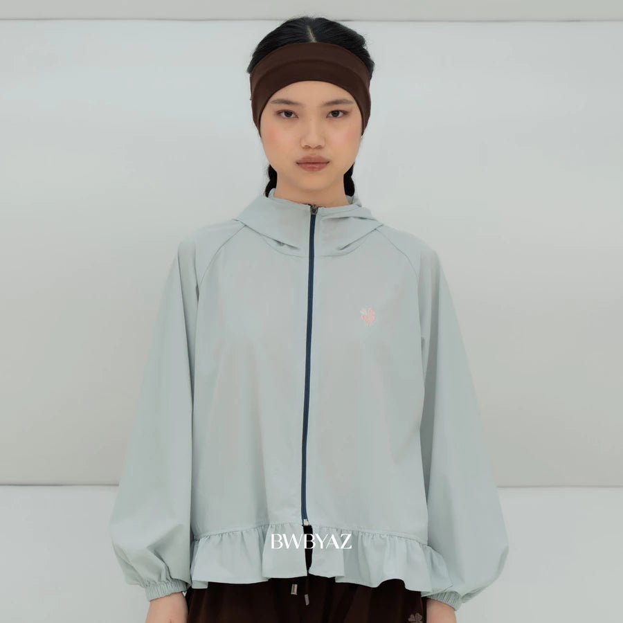 A model wearing a light awua blue hooded windbreaker jacket by BWBYAZ with a dark navy zipper, a small pink embroidered logo on the chest, and a ruffled hem. She is also wearing a dark brown headband and dark brown pants against a plain light gray studio background.