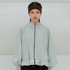 A model wearing a light awua blue hooded windbreaker jacket by BWBYAZ with a dark navy zipper, a small pink embroidered logo on the chest, and a ruffled hem. She is also wearing a dark brown headband and dark brown pants against a plain light gray studio background.