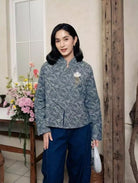 Woman wearing a patterned blouse by Haidee Orlin and blue pants standing indoors with flowers and a wooden door in the background.