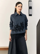 Woman wearing a navy blouse with floral embroidery by Studio Tui and black skirt against a white wall.