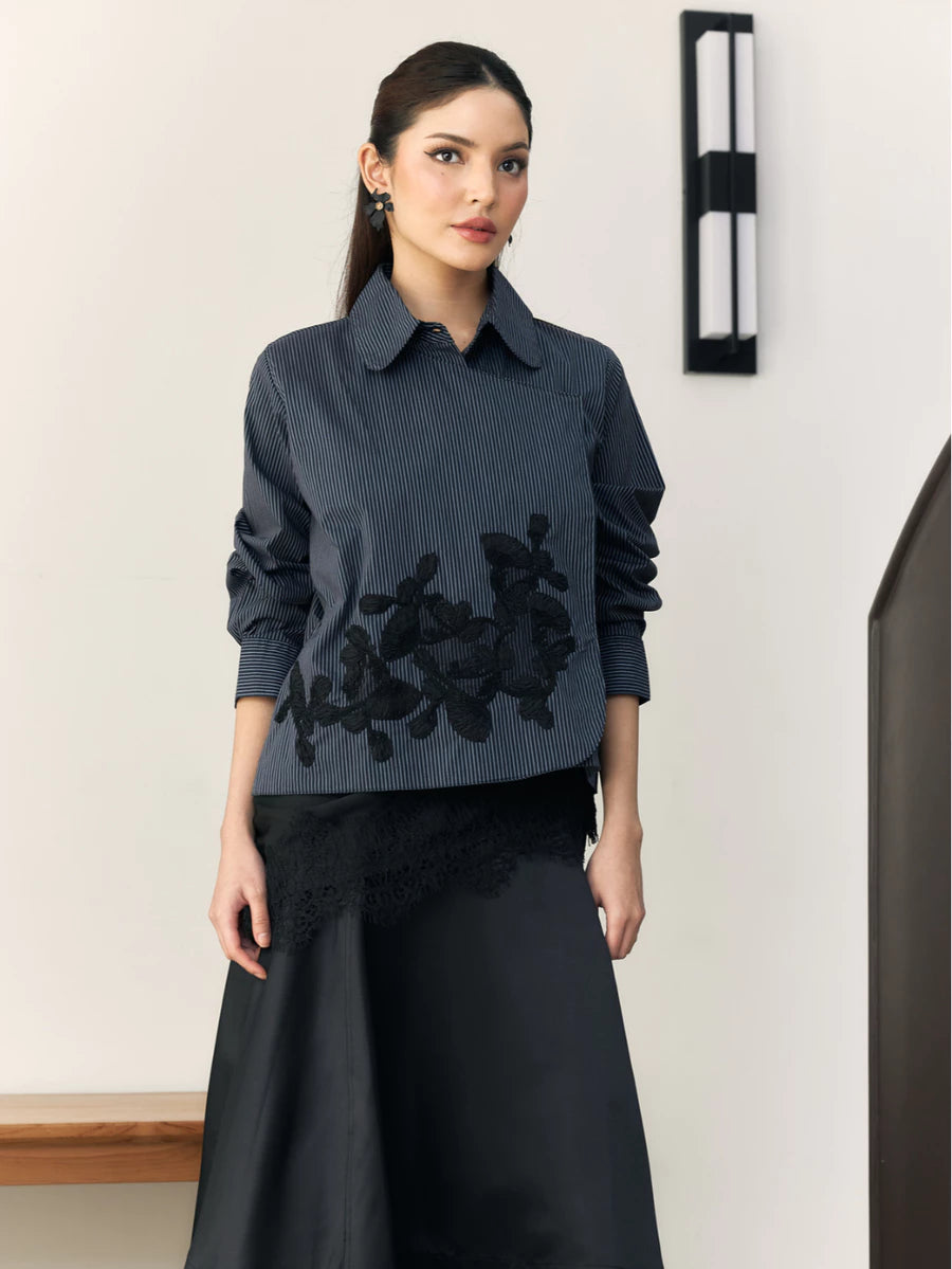 Woman wearing a navy blouse with floral embroidery by Studio Tui and black skirt against a white wall.