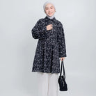 Woman wearing a black patterned tunic and white pants, holding a black bag on a white background