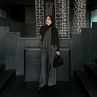Woman in a black jacket and dark grey pants by Renaette standing in a dark indoor setting with geometric light installations.