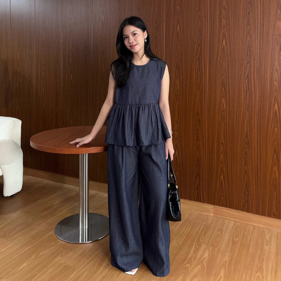 Woman in a dark denim top and pants by Renaette standing next to a wooden table against a wooden wall.