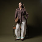 Woman in a brown asymetrical shirt from BWBYAZ and white pants holding a woven bag against a dark background