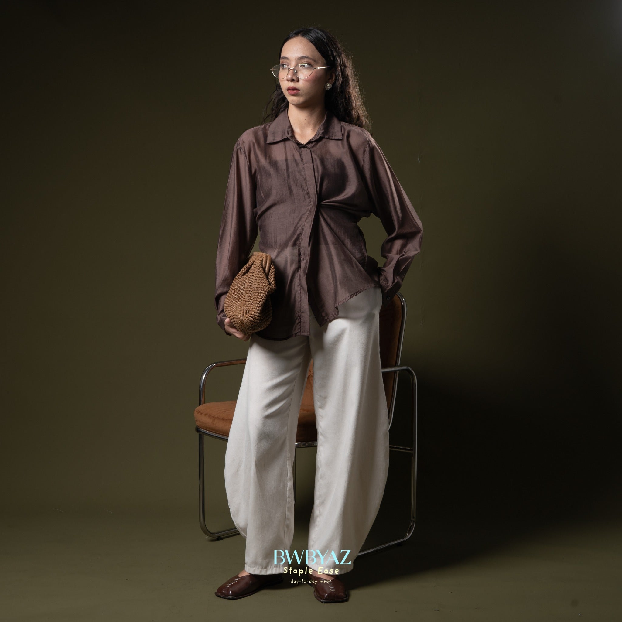 Woman in a brown asymetrical shirt from BWBYAZ and white pants holding a woven bag against a dark background