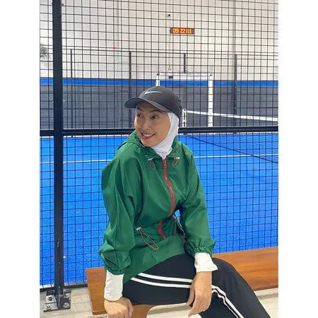 Person wearing a green track jacket by Abame and black cap sitting on a bench in an indoor sports facility.