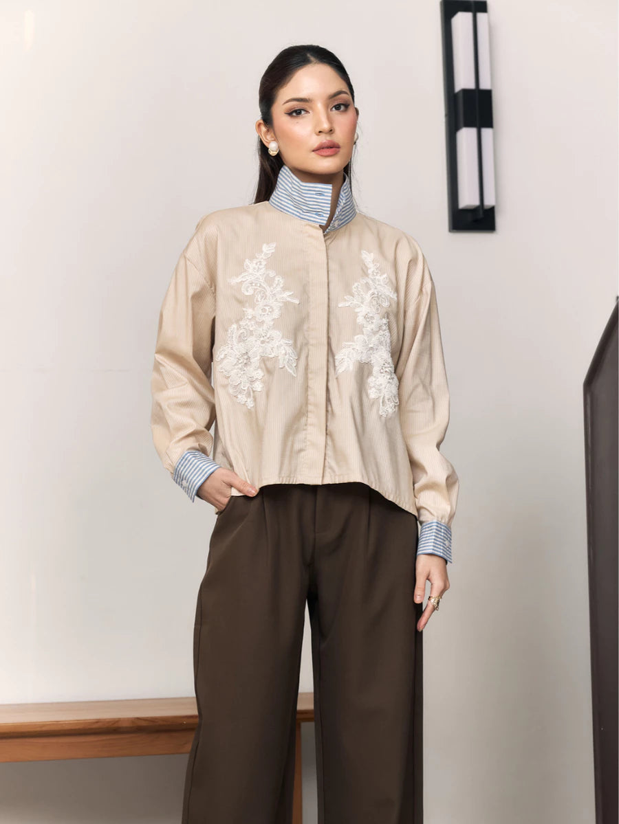 Woman wearing a beige shirt with floral embroidery by Studio Tui and brown pants against a plain background
