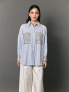 A woman modeling a light blue and white vertically striped button-down shirt by Studio Tui with decorative black-and-white patterned chest panels and striped cuffs, paired with wide-leg cream pants, against a dark gray studio background.