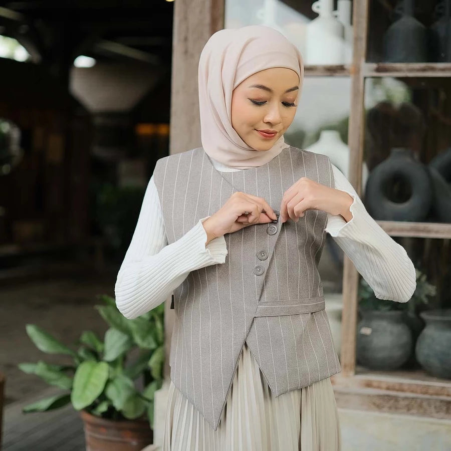 Woman wearing a gray pinstripe vest by Tunique over a white long-sleeve shirt, standing in an outdoor setting with plants.