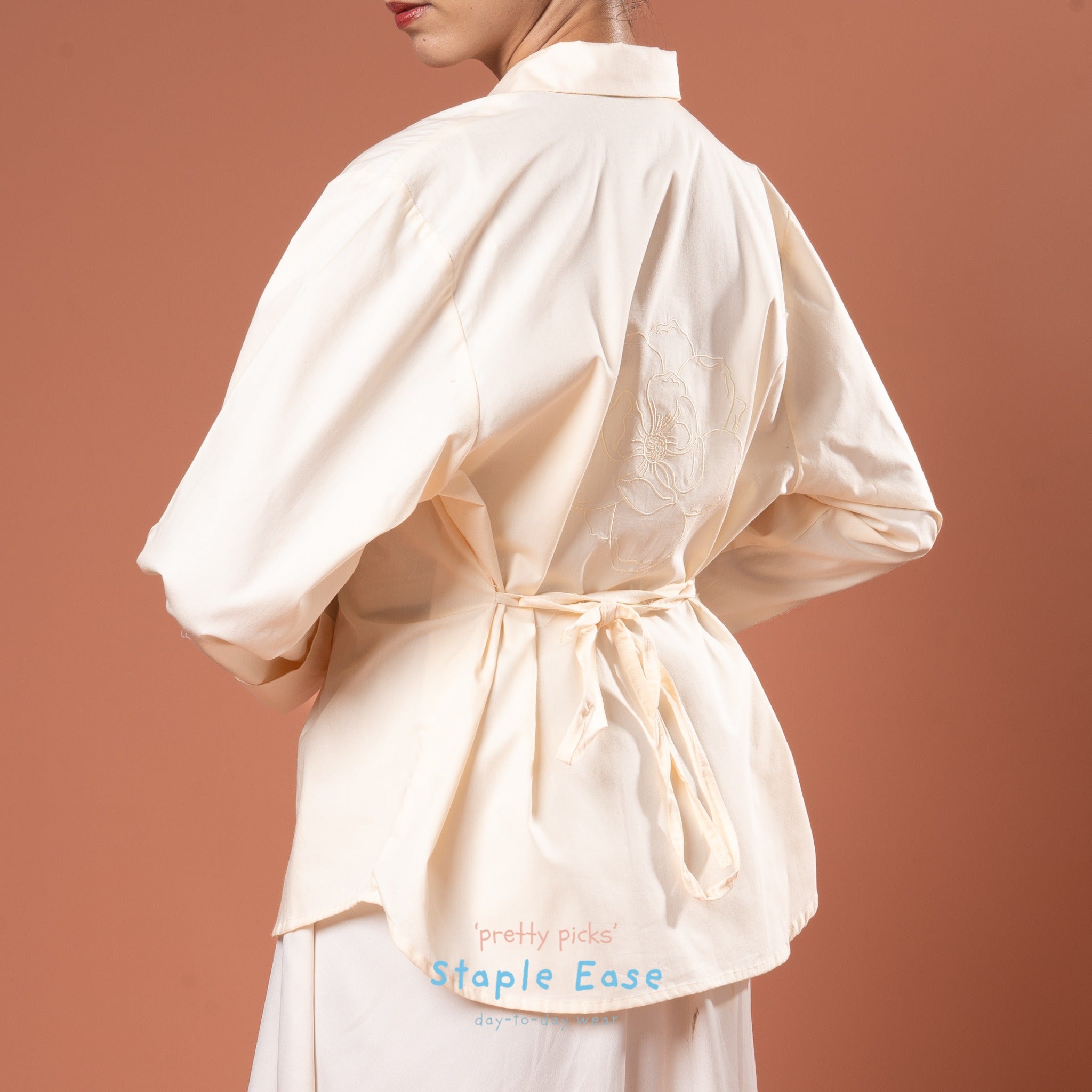 Person wearing a cream-colored blouse with a tie waist and floral embriodery on the back by BWBYAZ on a brown background