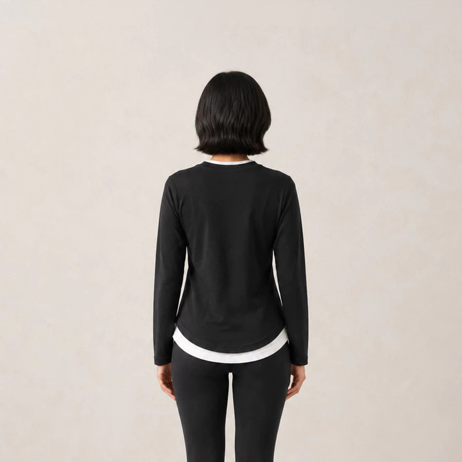 A woman viewed from behind wearing a black long-sleeve athleisure t-shirts by Jenna & Kaia with white contrast trim at the collar and hem, paired with black leggings, against a plain light beige background.