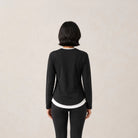 A woman viewed from behind wearing a black long-sleeve athleisure t-shirts by Jenna & Kaia with white contrast trim at the collar and hem, paired with black leggings, against a plain light beige background.