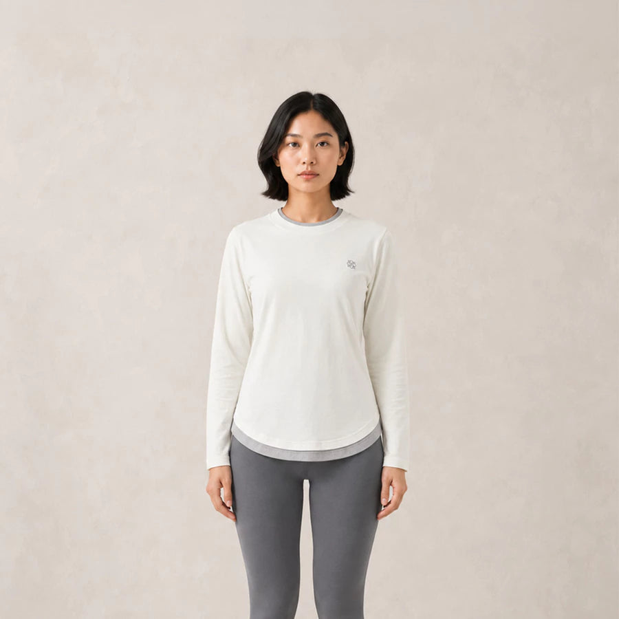 A woman modeling a white long-sleeve athletic top by Jenna & Kaia with a small embroidered logo on the left chest, paired with gray leggings, against a neutral beige textured background.