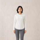 A woman modeling a white long-sleeve athletic top by Jenna & Kaia with a small embroidered logo on the left chest, paired with gray leggings, against a neutral beige textured background.