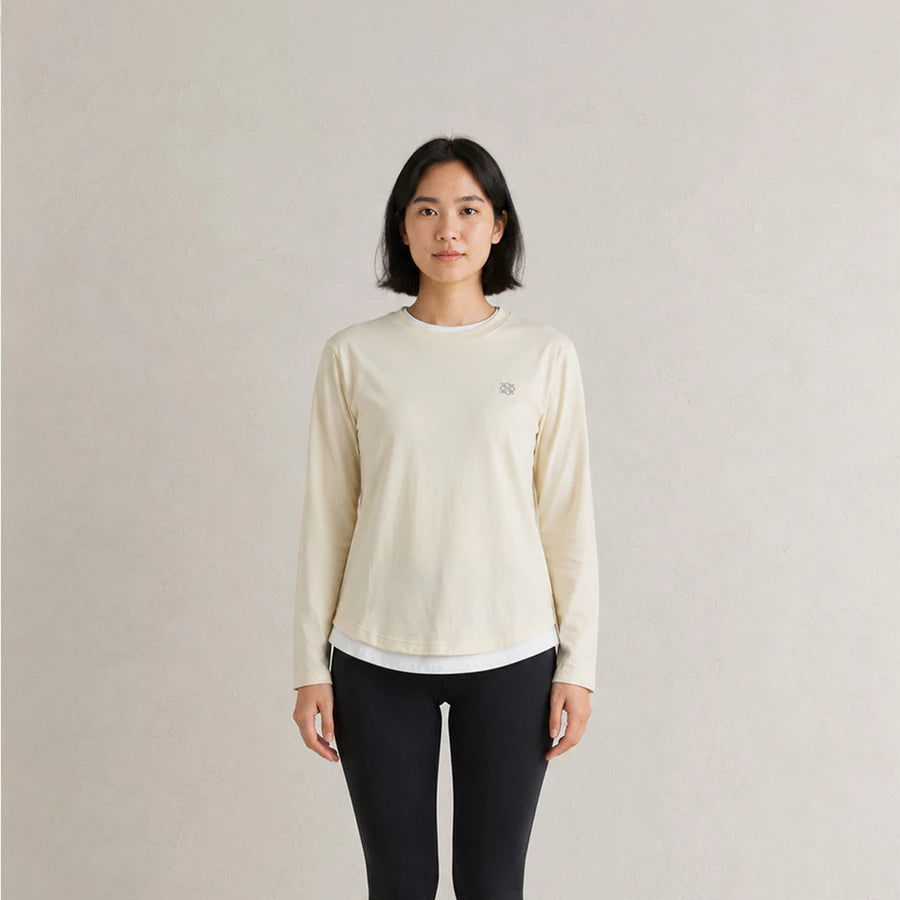 A woman stands against a light beige textured wall wearing a cream-colored long-sleeve athletic top by Jenna & Kaia with a small embroidered logo on the left chest, paired with black leggings.