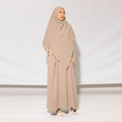A woman standing in a studio wearing a beige khadija abaya set by Lozy Hijab with a matching headscarf, designed for coverage and elegance in a neutral color.