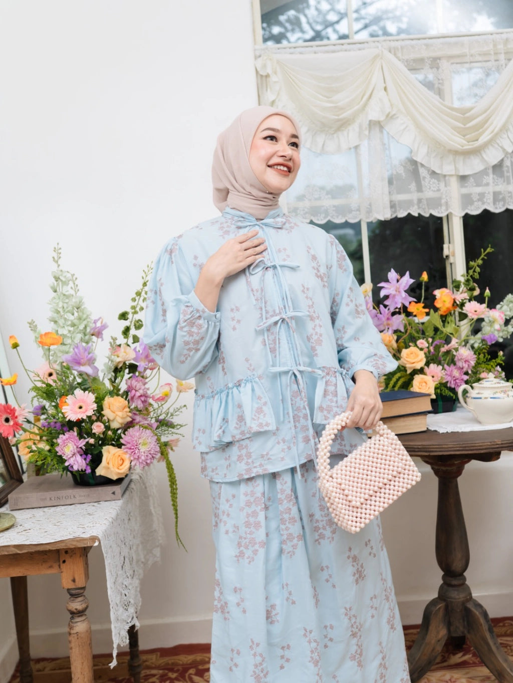 Woman in a light blue floral outfit by Haidee Orlin standing in a room with flowers and a table.