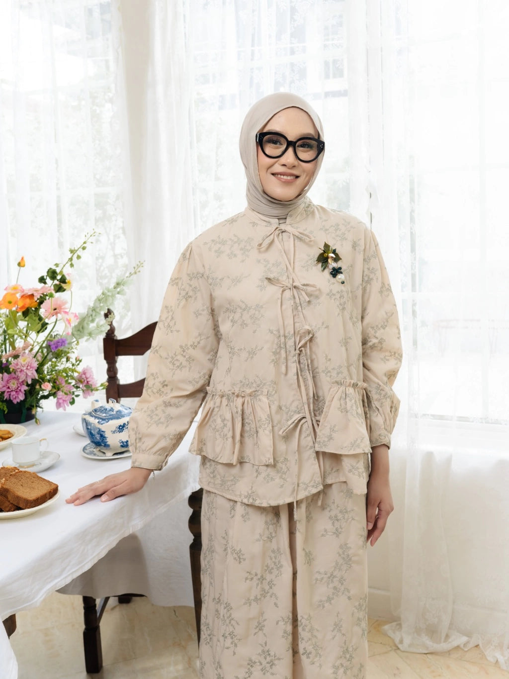 Person wearing a cream floral outfit by Haidee Orlin standing in a bright room with white curtains and a table with flowers.