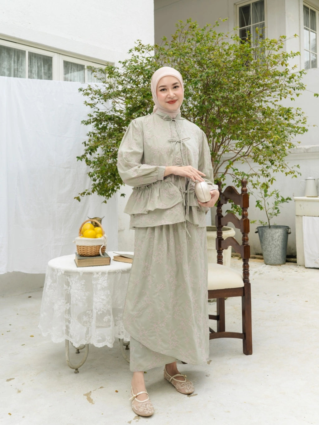 Woman in a light sage traditional outfit by Haidee Orlin standing in a bright room with plants and furniture.