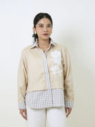A woman modeling a beige jacket by Studio Tui with white floral embroidery on the chest and white and gray gingham layering, paired with white pants, against a plain off-white background.