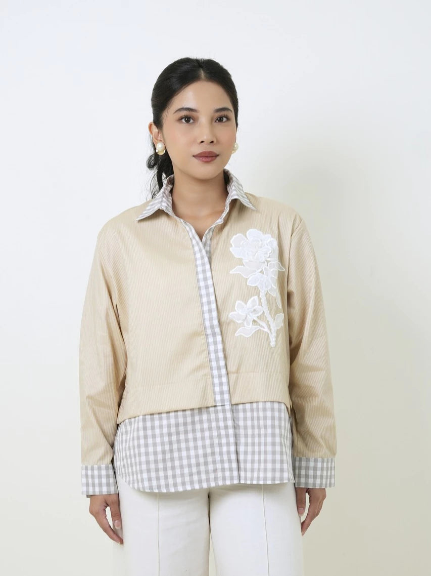 A woman modeling a beige jacket by Studio Tui with white floral embroidery on the chest and white and gray gingham layering, paired with white pants, against a plain off-white background.