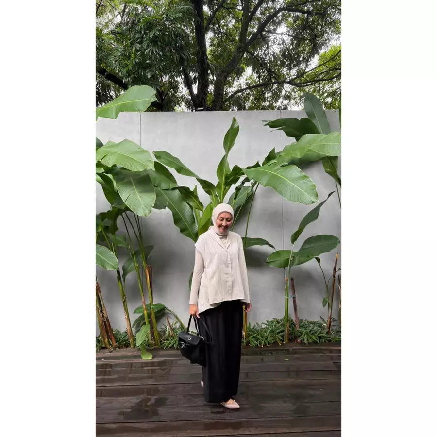 Woman in a beige vest by Abame over a beige shirt standing in front of a white wall with green plants