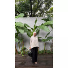 Woman in a beige vest by Abame over a beige shirt standing in front of a white wall with green plants
