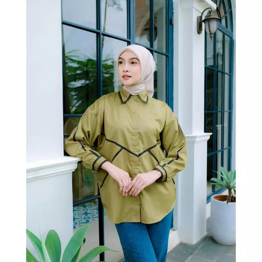 Woman wearing a green shirt by Abame and blue pants standing in front of a white building with glass doors.