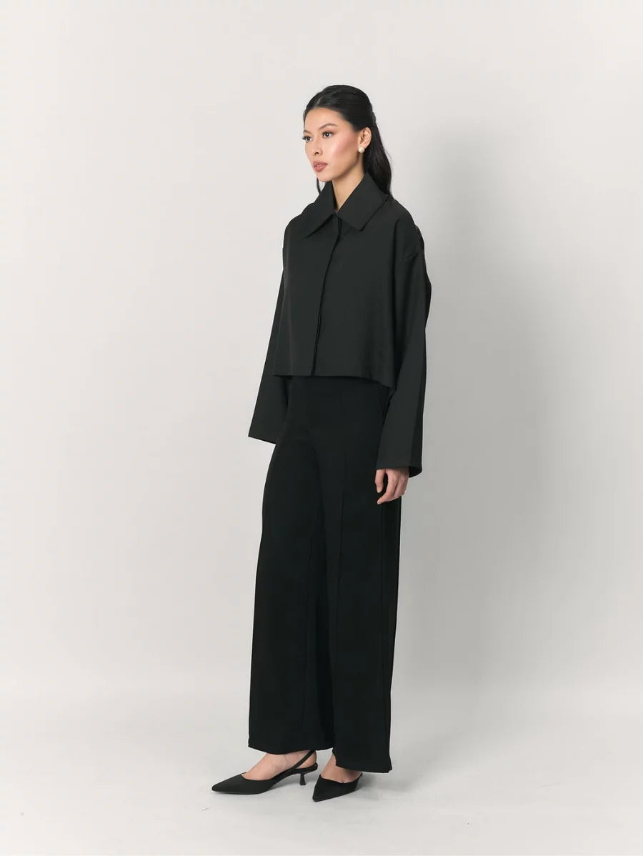 A model stands in profile wearing a black cropped long-sleeve jacket by Studio Tui with a wide collar, paired with black wide-leg trousers and black pointed-toe heels, against a light gray studio background.