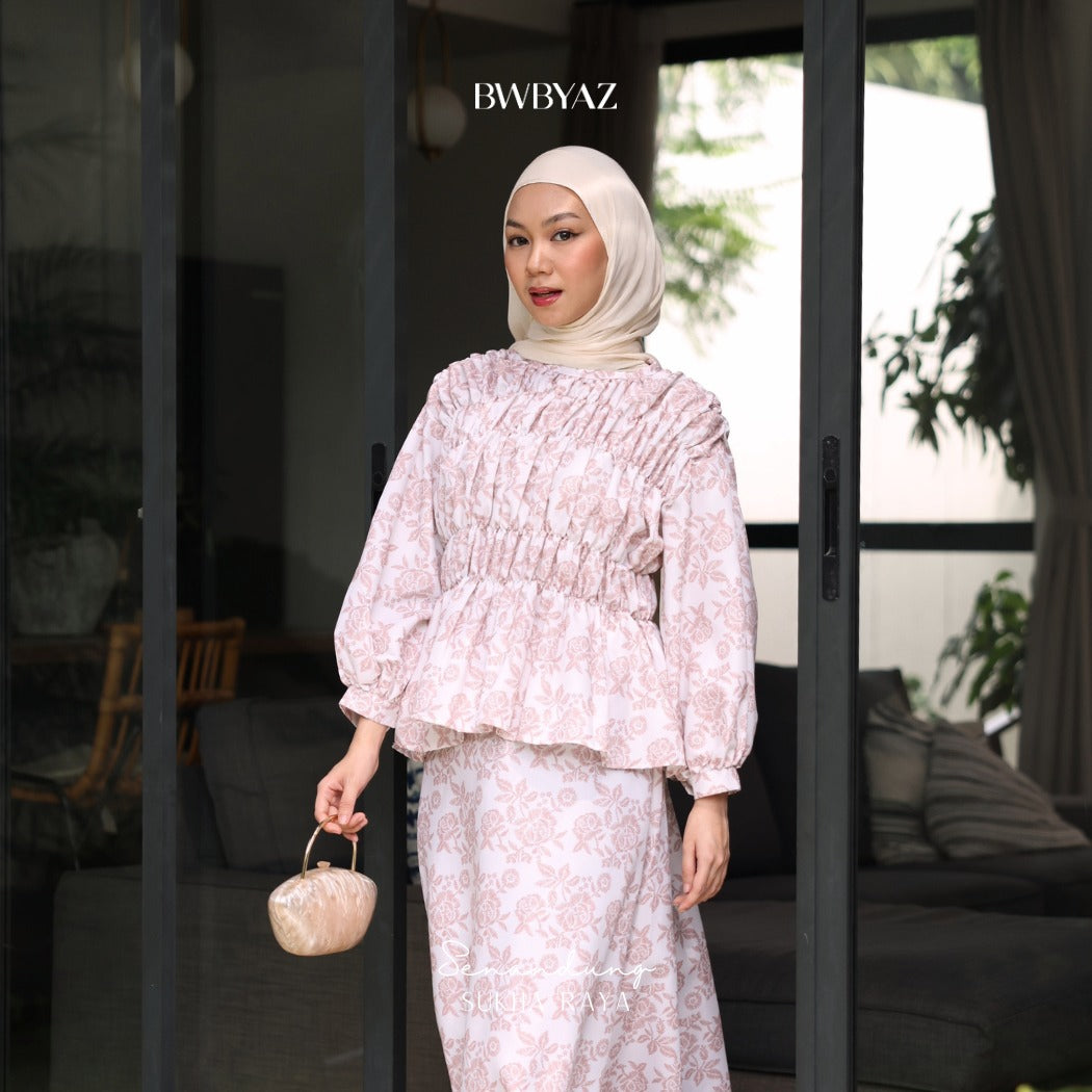A woman wearing a pink floral blouse with smocked details on the chest and waist from BWBYAZ, and a matching pink headscarf. She is holding a small purse and standing in front of a glass door.