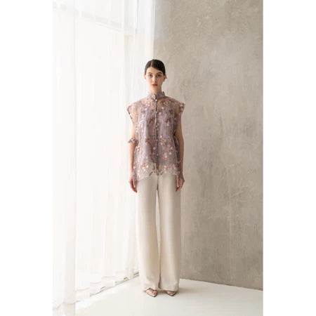 A woman is standing in a room wearing a lilac floral embroidered brocade vest with a Mandarin collar and light beige trousers.