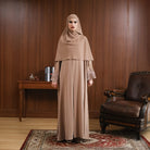Woman in a brown hijab and abaya by Tubita standing in a room with wooden walls and a leather chair.