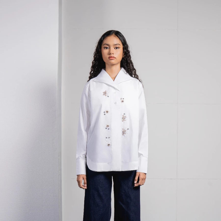 A model stands against a light gray tiled wall wearing a white long-sleeve button-up shirt by Jenna & Kaia with delicate floral embroidery on the front and dark navy wide-leg pants.