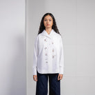 A model stands against a light gray tiled wall wearing a white long-sleeve button-up shirt by Jenna & Kaia with delicate floral embroidery on the front and dark navy wide-leg pants.