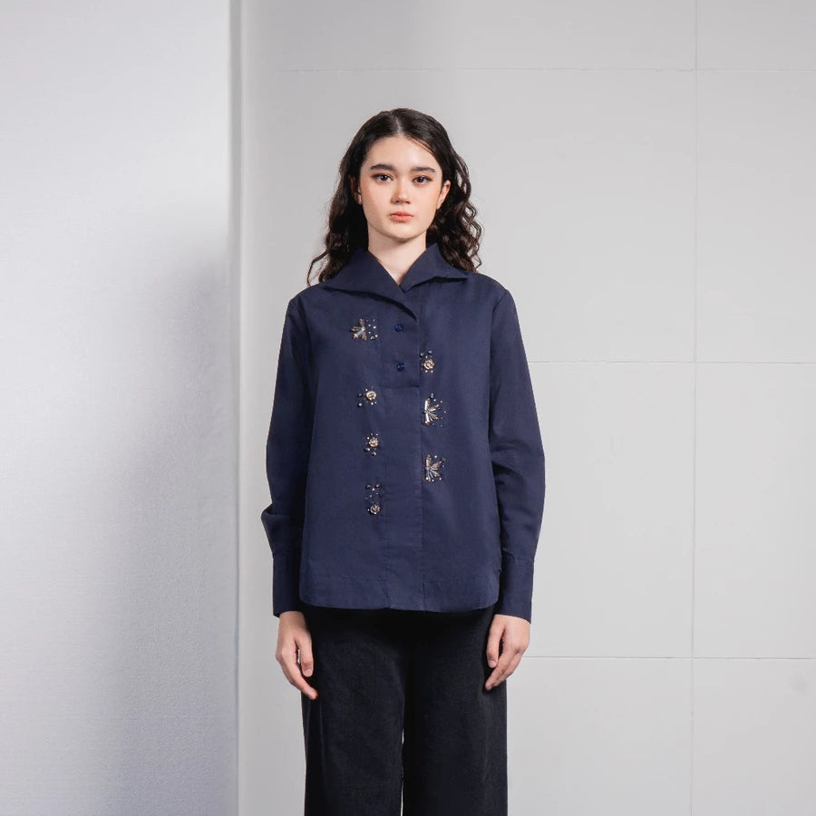A woman stands against a light gray tiled wall wearing a dark navy long-sleeve button-front shirt by Jenna & Kaia with small embroidered details and dark wide-leg pants.
