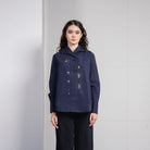 A woman stands against a light gray tiled wall wearing a dark navy long-sleeve button-front shirt by Jenna & Kaia with small embroidered details and dark wide-leg pants.